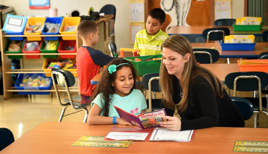 A teacher reads to a child while two students sit at a table behind them