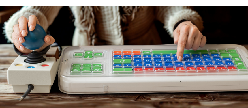 A close-up view of a person using assistive technology on a wooden desk, gripping a large blue joystick controller with their left hand and typing on a color-coded, high-contrast accessible keyboard with their right index finger.