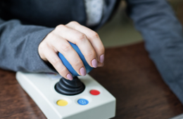 A close-up view of a person using assistive technology on a wooden desk, gripping a large blue joystick controller with their left hand.
