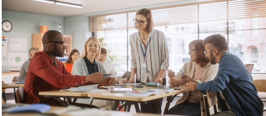 An image showing a diverse group of educators collaborating around a table in a bright classroom while a female leader with an ID badge stands to facilitate the discussion.