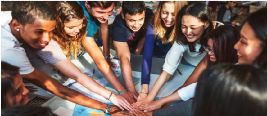 A diverse group of smiling young adults or students leaning over a table and placing their hands together in a central stack, symbolizing teamwork, unity, and collaboration.