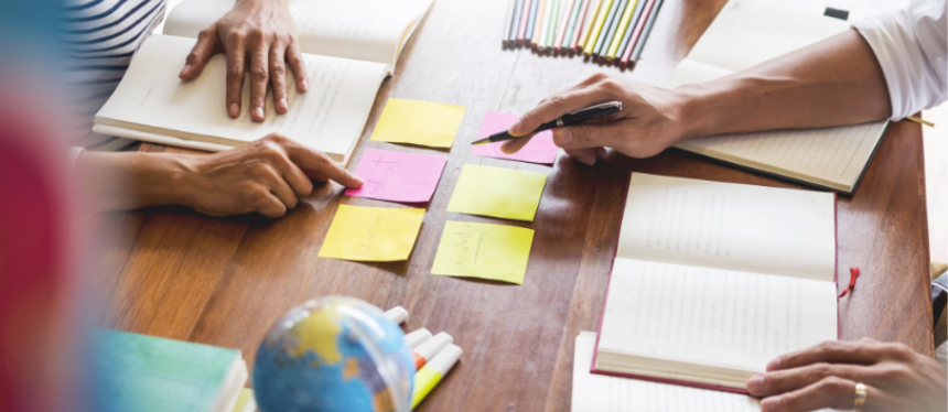 A top-down view of people collaborating at a wooden table, pointing at pink and yellow sticky notes arranged between open notebooks and study materials