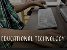 A student sitting at a wooden desk wearing a plaid shirt. One hand is on the computer mouse, while the other hand rests on the table next to the keyboard. The text reads "Educational Technology."