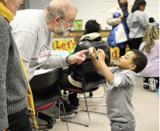 A side-profile photo of a man with glasses and a beard leaning down to interact with a young boy holding a small wooden block.  They are in a busy classroom or community workshop environment.