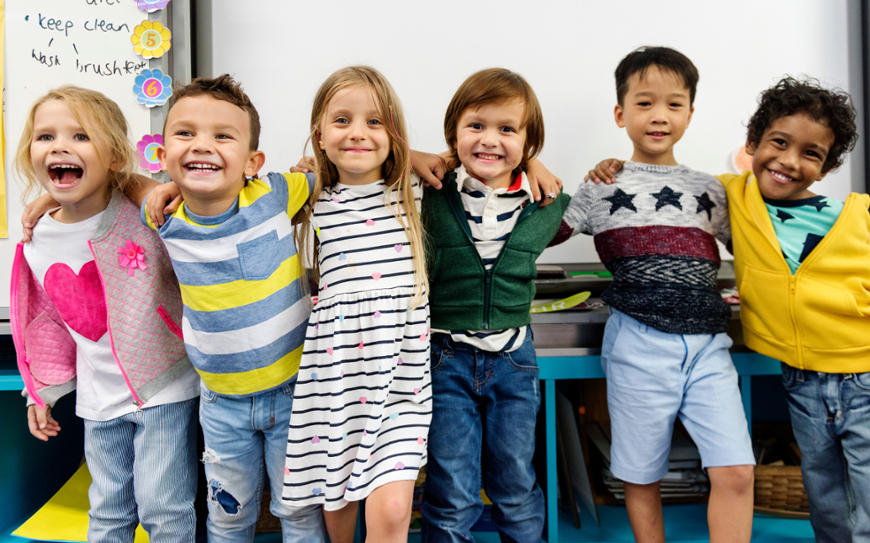 A joyful and diverse group of six young elementary school children standing in a row with their arms wrapped around each other's shoulders