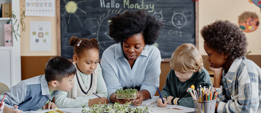 A female teacher and a diverse group of four elementary school children gathering around a desk to examine a small tray of green sprouts. A blackboard in the background reads "Hello Monday."