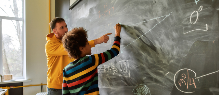 A side view of an adult teacher in a yellow hoodie guiding a young student wearing a striped sweater as they solve a geometry problem on a large chalkboard.