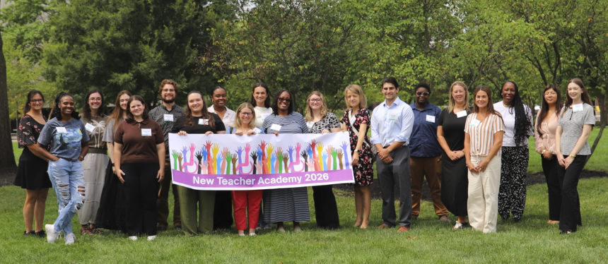 A large group of educators stands together on a grassy lawn, holding a banner that reads "New Teacher Academy 2026" featuring an illustration of colorful raised hands.