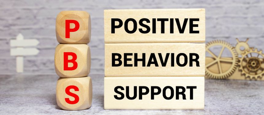 Wooden blocks arranged to spell out the acronym PBS. Three stacked wooden dice on the left display the red letters P, B, and S vertically. To their right, three stacked rectangular wooden blocks display the words "POSITIVE," "BEHAVIOR," and "SUPPORT" in bold black text.
