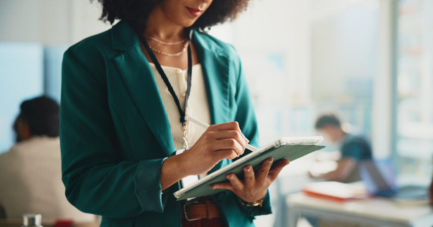 A close-up shot of an educator or professional woman with curly hair wearing a teal blazer and an ID badge lanyard. She is standing and focused on writing or drawing on a tablet with a white digital stylus, with a blurred classroom environment in the background.