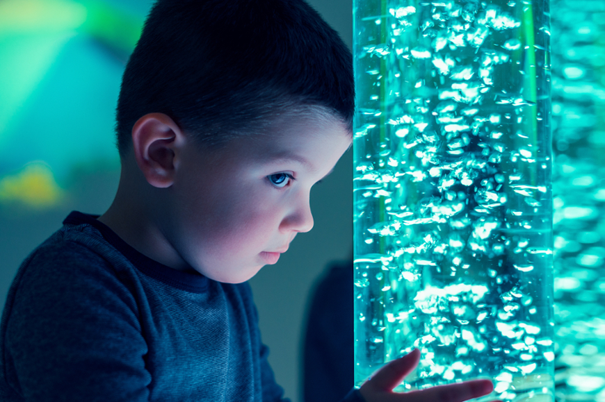 A close-up profile view of a young boy looking intently at a glowing teal sensory bubble tube in a dimly lit room.