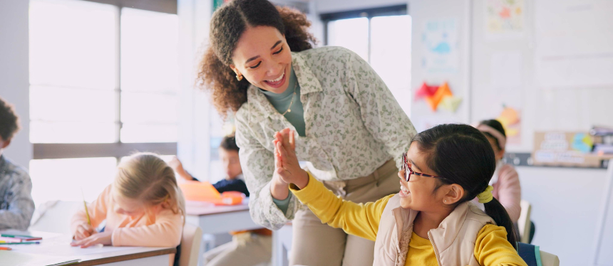 A smiling female educator leans over a classroom desk to give a young girl with glasses a high-five during a lesson