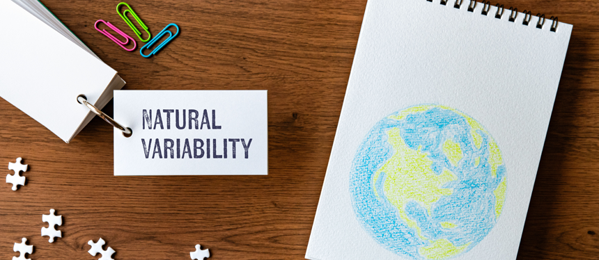 A wood background with colored paper clips, a ring of white flashcard including the text "natural variability," white puzzle pieces and a white notepad with an illustrated green and blue globe centered on the cover page.